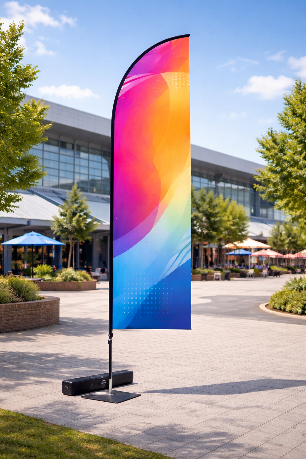 Large gradient feather flag with flat bottom edge on black base displayed in outdoor commercial plaza with clear blue sky